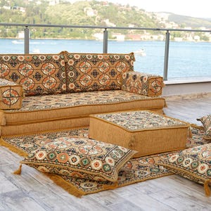 May include: A low-profile, modular seating set with a patterned design in shades of brown, beige, and orange. The set includes a sofa, ottoman, rug, and several throw pillows. The pieces are arranged on a wooden deck with a scenic waterfront view.