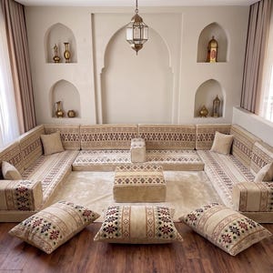 May include: A U-shaped floor seating arrangement featuring beige cushions with intricate geometric patterns in red, brown, and black. The set includes backrests, armrests, a central square ottoman, and three large floor pillows on a wooden floor.