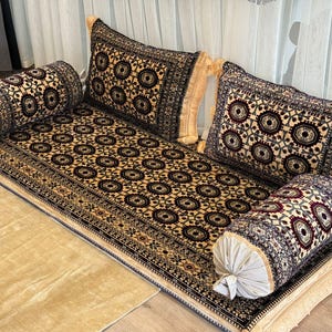 May include: A decorative floor seating set featuring a patterned rug, two bolster pillows, and two square pillows. The rug and pillows have a beige background with a repeating circular design in dark brown, burgundy, and cream. The set has a fringed edge.