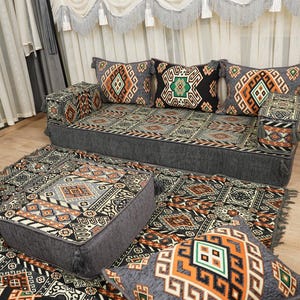 Può includere: Un set di sedute a pavimento grigio con cuscini fantasia e un tappeto abbinato. I cuscini presentano disegni geometrici arancioni, neri e verdi. Il set comprende un divano, due pouf e diversi cuscini.