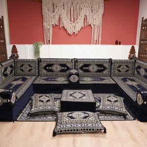 May include: A large, U-shaped sofa with a low coffee table and floor cushions. The furniture is navy blue with a detailed, geometric pattern in gold and cream. The room has wooden room dividers and a macrame wall hanging.