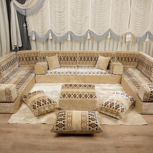 May include: A beige U-shaped sofa with patterned cushions and an ottoman. The sofa has decorative pillows and sits on a rug. The curtains and wall decor complement the sofa's colour scheme. The sofa is approximately 300cm x 300cm.