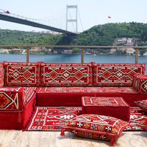 May include: A red sectional sofa with patterned cushions and a matching rug. The sofa is arranged on a patio overlooking a body of water and a bridge. The cushions and rug feature a geometric design.