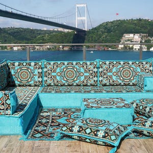 May include: A turquoise corner sofa with patterned cushions and a matching rug. The cushions have a geometric design in black, brown, and cream. The sofa is on a wooden deck with a waterfront view.