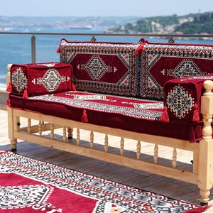 Puede incluir: Un sofá de exterior de madera con un cojín y almohadas estampadas en rojo. El sofá tiene una estructura de madera clara y está colocado en una terraza con una alfombra roja a juego. Los cojines y almohadas tienen un diseño geométrico con borlas.