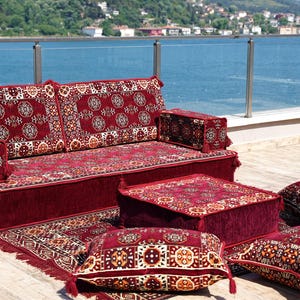 May include: An outdoor seating arrangement comprising a burgundy sofa, ottoman, and cushions. The set displays an intricate pattern in red, cream, and gold. The scene is set against a backdrop of a blue sea and sky. The sofa is approximately 180 cm long.