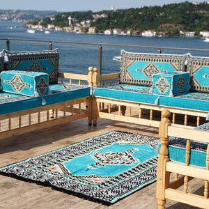 May include: Outdoor seating set with light wood frames and turquoise cushions. The set includes two sofas, a loveseat, and a matching rug. The cushions feature a patterned design in shades of turquoise, black, and cream. The furniture is arranged on a wooden deck overlooking a body of water.