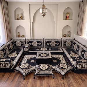 May include: A modular floor sofa set featuring black fabric with intricate beige and brown geometric patterns. The set includes long back cushions, a central square ottoman, and three floor pillows, all matching in design and color scheme.