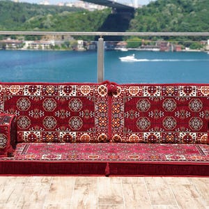 Puede incluir: Un sofá de suelo rojo con intrincados diseños estampados. El sofá tiene un perfil bajo con una base rectangular y dos reposabrazos. El fondo presenta una vista panorámica del agua y un puente.