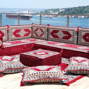 Puede incluir: Un juego de sofás modular rojo con cojines estampados y una alfombra a juego. El conjunto incluye un sofá esquinero, otomanas cuadradas y cojines decorativos con borlas. El diseño presenta patrones geométricos en tonos rojos, blancos y negros.