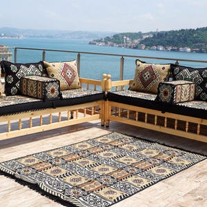 May include: Two wooden benches with black cushions and patterned pillows. A matching rug lies in front. The benches are on a patio overlooking a body of water and a distant shoreline. The pillows have red, black, and beige geometric designs.