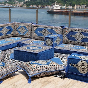 Puede incluir: Un conjunto modular de asientos para exteriores con un diseño azul y estampado. El conjunto incluye cojines, otomanas y una alfombra, todos con un patrón geométrico en tonos azules, beige y negro. El conjunto está dispuesto sobre una terraza de madera con vistas al agua.