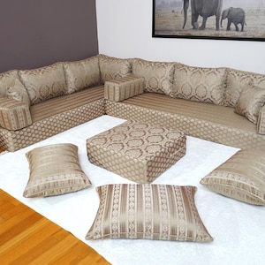 May include: A beige sectional sofa with patterned cushions and a matching square ottoman. The sofa is arranged in an L-shape and sits on a white rug. Several decorative pillows are arranged on the rug. The room has hardwood floors and a framed picture on the wall.