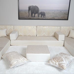 May include: A cream and beige modular sofa with patterned cushions and a matching square ottoman. Decorative pillows and a framed elephant print create a cohesive living room design. The sofa is designed for comfort.
