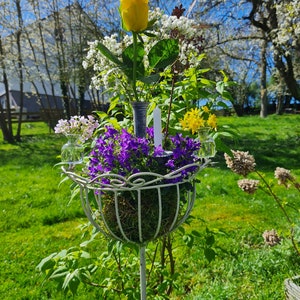 Könnte beinhalten: Ein weißer Pflanzenständer aus Metall mit einem kugelförmigen Korb, gefüllt mit lila Blumen, Moos und anderem Grün. Eine gelbe Rose und andere Blumen sind darüber angeordnet. Eine weiße Kerze befindet sich in der Mitte.