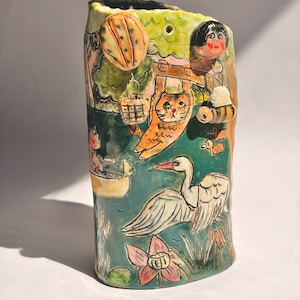 May include: A cylindrical ceramic vase featuring a hand-painted scene with a white swan, a person in a yellow boat, a hanging orange cat, a hot air balloon, a bee, and a small figure. The surface is decorated with green, blue, and pink tones.