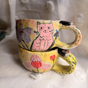 May include: Two stacked ceramic mugs with hand-painted designs. The top mug features a pink cat and purple flowers on a yellow background. The bottom mug displays a large red and pink flower. Both mugs have yellow handles with black dots.