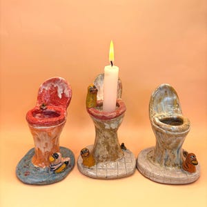 May include: Three ceramic toilet-shaped candle holders in red, beige, and blue. One holds a lit white candle. Each has a small, whimsical creature attached to the base. The candle holders are decorative.