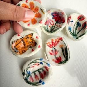 May include: A collection of small, hand-painted ceramic dishes. Each dish features a unique design, including flowers, a tiger, and abstract floral patterns. The color palette includes orange, red, blue, and green on a speckled white background.