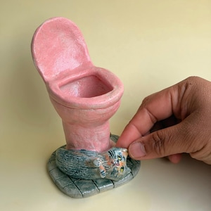May include: A pink ceramic toilet-shaped sculpture featuring a small, textured grey and orange cat lying on the base. The base is designed with a grid pattern resembling tiled flooring, and the toilet has an upright lid and a hollow bowl.