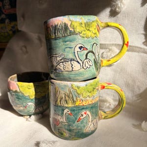 May include: Two handmade ceramic mugs and one small bowl featuring hand-painted scenes of white swans swimming in a blue pond with green foliage. The mugs have bright yellow handles with small red floral accents and a glossy glazed finish.