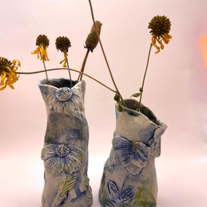 Pode incluir: Dois vasos de cerâmica com um design floral azul e branco. Os vasos são feitos à mão e têm um acabamento rústico e texturizado. Eles estão cheios de flores secas.