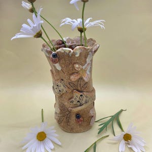 Könnte beinhalten: Eine handgefertigte, hellbraune Keramikvase mit strukturierter Oberfläche, mit geprägten Blumen- und Tiermotiven. Die Vase enthält weiße Gänseblümchen mit gelben Zentren. Ein Marienkäfer ist ebenfalls auf der Vase zu sehen. Die Vase ist ein dekorativer Artikel.