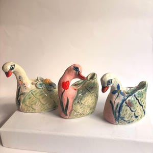 May include: Three ceramic swan-shaped planters or candle holders. The left swan is cream with a blue flower, the centre is pink with a red tulip, and the right is white and pink with a blue flower. Each features textured wings and orange beaks.