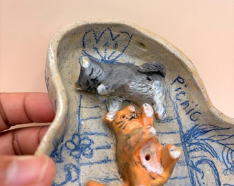May include: A ceramic dish with a light beige base, featuring blue sketched designs and the word "Picnic". Two small cat figurines are inside: a gray cat lying down and an orange cat on its back.