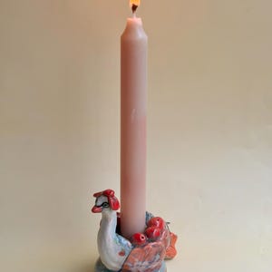 May include: A tall, light pink taper candle with a lit flame sits inside a ceramic candle holder shaped like a swan. The swan figure features a white neck, a red beak, and a body decorated with small red apple-like shapes and blue accents.