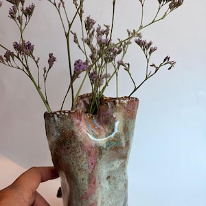 May include: A hand-sculpted ceramic vase with an irregular, organic rim and a glossy, mottled glaze in shades of sage green, dusty pink, and brown. The vase contains a cluster of dried purple statice flowers with thin, branching green stems.