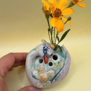 May include: A handcrafted ceramic bud vase with a swan design, in white, blue, and green. The vase holds bright yellow flowers. The vase has three holes for stems and a flower design. The vase is approximately 10 cm in diameter.