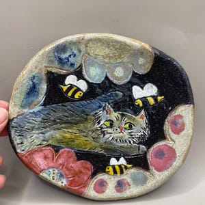May include: Handmade ceramic plate with a whimsical design. A grey and white cat is surrounded by three yellow and black bees, red flowers, and blue and white clouds. The plate has a black background and a textured, irregular edge.