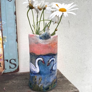 May include: A hand-painted cylindrical vase featuring a swan and water scene, holding white daisies with yellow centres. The vase has a pink and blue colour scheme, with a small bird figurine at the top. The vase is on a wooden surface.