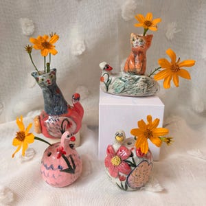 May include: Four quirky ceramic vases, each shaped like an animal, hold bright orange flowers. The vases include a cat, a duck, and a bird, all with colourful painted details. The vases are displayed on a white surface.