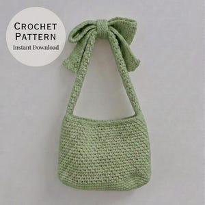 Peut inclure: Un sac au crochet vert sauge clair doté d'une bandoulière unique ornée d'un grand nœud proéminent sur le dessus. Le texte CROCHET PATTERN Instant Download est imprimé en noir sur un fond circulaire blanc dans le coin supérieur gauche.