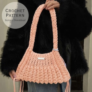 May include: A peach-colored crochet handbag featuring a thick braided shoulder strap and a rectangular silver-toned metal tag on the front. The bag has a textured, ribbed knit construction and includes a digital overlay text reading CROCHET PATTERN Instant Download.
