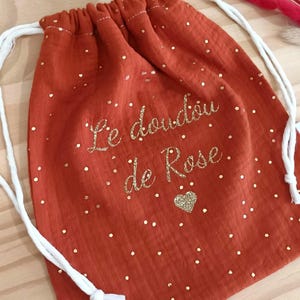 Peut inclure: Un sac à cordon de couleur rouille avec des liens blancs et un motif à paillettes dorées composé de petits points et d'un cœur. Le texte "Le doudou de Rose" est imprimé en paillettes dorées sur le devant du sac en tissu texturé.