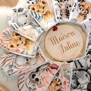 Puede incluir: Un conjunto de 10 mordedores de tela para bebés con diferentes diseños de animales, incluyendo un león, un elefante, un mono y un guepardo. Los mordedores están dispuestos en círculo alrededor de un letrero de madera que dice "Maison Lutine".