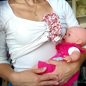 Op de afbeelding: Een wit geribbeld shirt met een bloemenprint borstvoedingshoes. Een gouden hartvormige hangerketting is zichtbaar. Een pop in een roze jurk wordt in de armen gehouden.