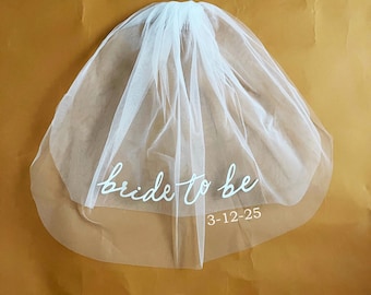 May include: White bridal veil with the text "bride to be" and the date "3-12-25" written on it.