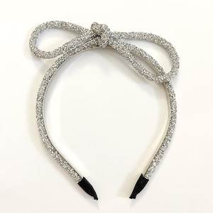 May include: A silver headband with a sparkly bow. The headband is made of a thin metal band with a silver glitter finish. The bow is made of the same material and is tied in a simple knot.
