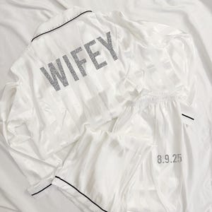 May include: White satin pajama set with a black trim. The top has the word "WIFEY" in silver glitter on the back. The pants have the number "8.9.25" in silver glitter on the leg.