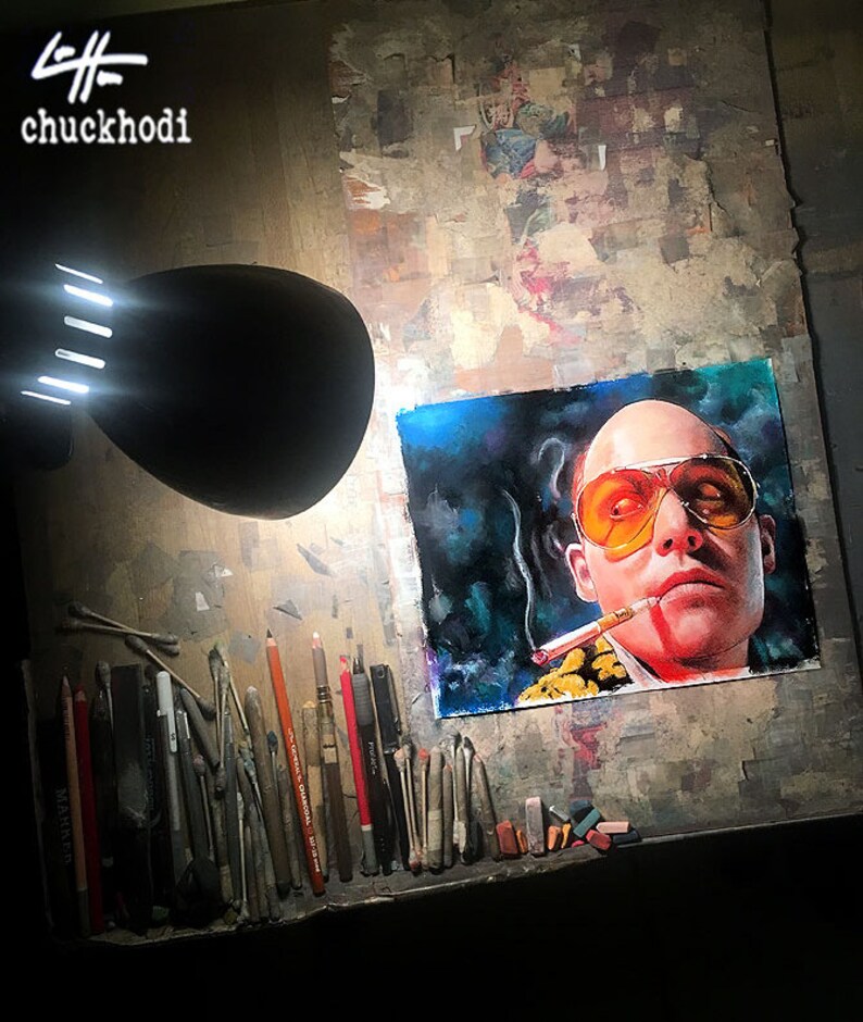 May include: A close-up of a drawing of a man with a cigarette in his mouth, wearing sunglasses and a yellow shirt. The drawing is on a white background and is surrounded by art supplies, including pencils, erasers, and cotton swabs.