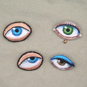 May include: Four embroidered eye patches with detailed stitching. Two patches feature blue eyes, one with a green eye, and another with a brown eye. Each eye is outlined with a black beaded border, and the background is a neutral beige fabric.