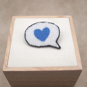 May include: A beaded brooch shaped like a speech bubble with a white background and a blue heart in the centre. The speech bubble is outlined with a thin border of dark grey beads, creating a distinct contrast against the white and blue interior.