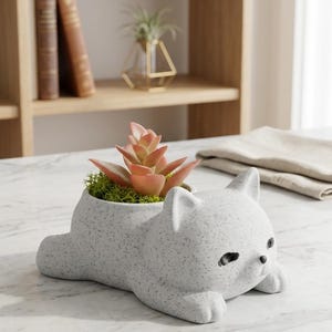 May include: A white ceramic planter shaped like a reclining cat with a speckled gray texture. The cat has small black eyes and a nose. A pink succulent plant with green moss is nestled in the opening on the cat's back, creating a decorative piece.