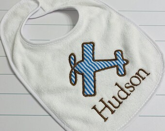 May include: White terrycloth baby bib with a blue and white striped airplane design and the name "Hudson" embroidered in brown thread.
