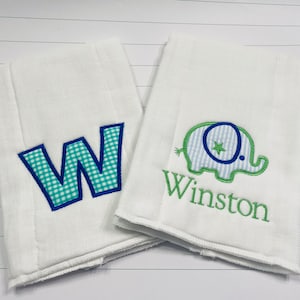 May include: Two white burp cloths with embroidered designs. One cloth features a blue and green checkered letter "W" and the other features a green elephant with a blue and white checkered body and the name "Winston" embroidered below.