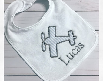 May include: White baby bib with an embroidered airplane design in gray and light blue stripes. The name "Lucas" is embroidered below the airplane. The bib has a rounded shape with a snap closure at the neck.